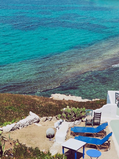 The scene features a picturesque coastal setting with vibrant turquoise waters meeting the land. A patio area with blue lounge chairs, a small table, and a couple of black chairs is visible in the foreground. The area is bordered by white walls and greenery, including cactus plants. Rocks and the clear sea are prominent features, creating a serene, inviting atmosphere.