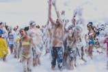 A group of people are enjoying a foam party, with laughter and playful expressions. Many are covered in white foam, and some are wearing swimsuits. The atmosphere is energetic, with arms raised and foam flying in the air.