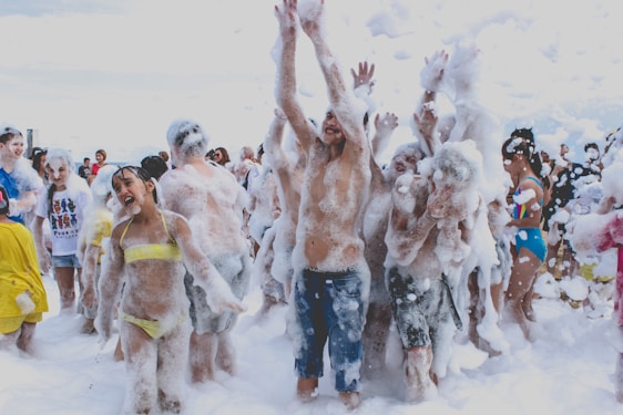 A group of people are enjoying a foam party, with laughter and playful expressions. Many are covered in white foam, and some are wearing swimsuits. The atmosphere is energetic, with arms raised and foam flying in the air.