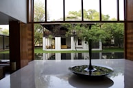A modern interior space with large windows showcasing a lush garden and traditional wooden house in the background. The room features a polished floor and a central circular water feature with plants.