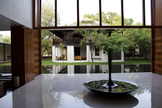 A modern interior space with large windows showcasing a lush garden and traditional wooden house in the background. The room features a polished floor and a central circular water feature with plants.
