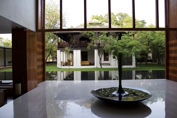 A modern interior space with large windows showcasing a lush garden and traditional wooden house in the background. The room features a polished floor and a central circular water feature with plants.