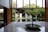 A modern interior space with large windows showcasing a lush garden and traditional wooden house in the background. The room features a polished floor and a central circular water feature with plants.