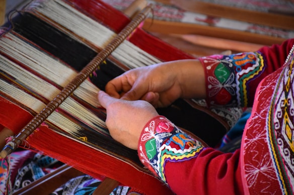 A person is engaged in traditional weaving, using a loom to create a patterned textile. The weaver's hands are in focus, manipulating threads of various colors including red, black, and white. The intricate designs on the fabric and on the person's clothing suggest a cultural or ethnic significance.
