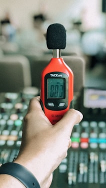 A hand holds a red sound level meter with a digital display reading 00.7. The background is blurred, suggesting an indoor environment, possibly with audio equipment nearby.