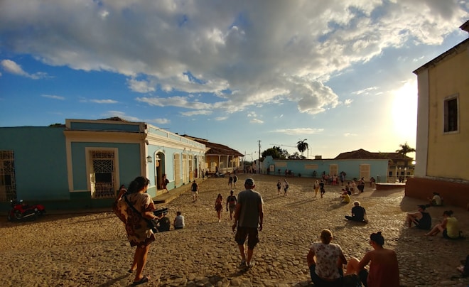 A sunny street scene in a colonial town with cobblestone pathways and colorful buildings. People are casually strolling, sitting, and enjoying the warm afternoon light with a few clouds scattered across the blue sky.