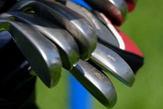 Close-up view of several golf clubs with metal heads, arranged in parallel. The background includes shades of green and hints of blue and red colors.