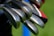 Close-up view of several golf clubs with metal heads, arranged in parallel. The background includes shades of green and hints of blue and red colors.