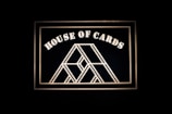 A geometric design resembling a house of cards is framed against a dark background. Bold, white lettering spells out 'House of Cards' above the central illustration.