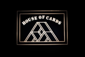 A geometric design resembling a house of cards is framed against a dark background. Bold, white lettering spells out 'House of Cards' above the central illustration.