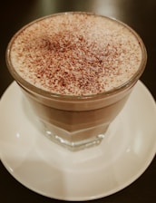 A glass mug filled with a frothy coffee beverage, topped with a layer of cream and sprinkled with cocoa or chocolate powder. It rests on a white saucer with a dark tabletop underneath.