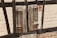 A close-up of a weathered wooden window shutter mounted on a rustic wall. The wall is partially constructed from dark wooden beams and light-colored bricks. The shutter has a rough, aged texture with visible knots and cracks in the wood.