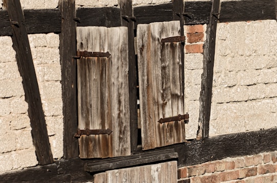 A close-up of a weathered wooden window shutter mounted on a rustic wall. The wall is partially constructed from dark wooden beams and light-colored bricks. The shutter has a rough, aged texture with visible knots and cracks in the wood.