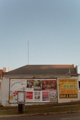 A single-story building with a corrugated metal roof serves as a billboard space for numerous posters advertising various events and products. Among them are concert posters for musical acts such as U2 and Boy & Bear, as well as promotional material for Australian hams. The building is situated along a street with a small grassy verge in front of it. The sky is clear with a faint blue color.