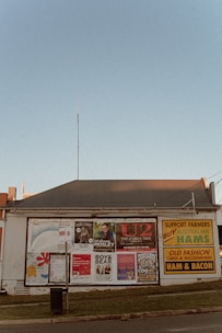 A single-story building with a corrugated metal roof serves as a billboard space for numerous posters advertising various events and products. Among them are concert posters for musical acts such as U2 and Boy & Bear, as well as promotional material for Australian hams. The building is situated along a street with a small grassy verge in front of it. The sky is clear with a faint blue color.