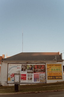 A single-story building with a corrugated metal roof serves as a billboard space for numerous posters advertising various events and products. Among them are concert posters for musical acts such as U2 and Boy & Bear, as well as promotional material for Australian hams. The building is situated along a street with a small grassy verge in front of it. The sky is clear with a faint blue color.