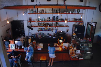 A cozy cafe with vintage decor featuring shelves stocked with various bottles and glasses. Several people are seated at the counter and tables, engaged in activities like reading and working on a laptop. Warm lighting from hanging bulbs adds to the inviting atmosphere.