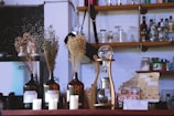 A cozy cafe scene featuring a variety of items on a counter. Brown glass bottles hold dried flowers, and there are white candles in front of the bottles. A unique coffee brewing device with a wooden frame and glass components sits prominently. In the background, shelves display various bottles, jars, and glassware, creating a rustic and eclectic atmosphere.