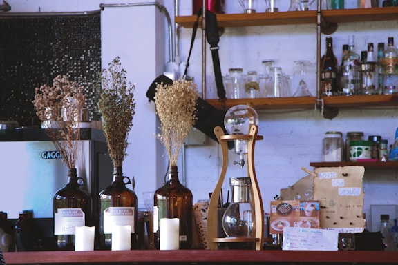 A cozy cafe scene featuring a variety of items on a counter. Brown glass bottles hold dried flowers, and there are white candles in front of the bottles. A unique coffee brewing device with a wooden frame and glass components sits prominently. In the background, shelves display various bottles, jars, and glassware, creating a rustic and eclectic atmosphere.