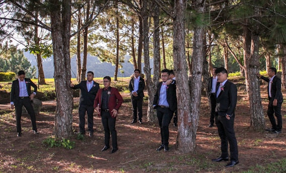 A group of people wearing formal attire, including suits and ties, stand in a wooded area among tall pine trees. The light filtering through the branches creates a pattern of shadows on the ground.
