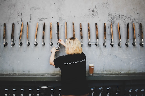 A person with blonde hair, wearing a black shirt with the words 'Pick Up A Hitchhiker', is standing at a bar with numerous wooden beer taps lined up against a textured gray wall. A glass filled with amber-colored beer sits on the counter beside them.