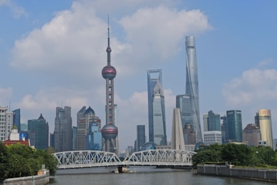 A vibrant cityscape features a modern skyline with iconic skyscrapers, including a distinctive tower with spherical structures. A river runs in the foreground, crossed by a large bridge, while the scene is bathed in daylight with scattered clouds in the sky.