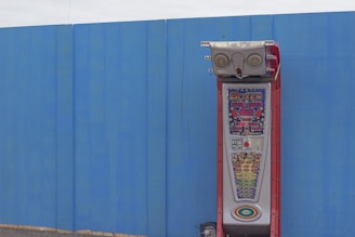 A boxing arcade machine stands against a large blue wall. The machine has a red structure with digital displays and colorful game indicators. It appears to be an outdoor setup, placed on a pavement.