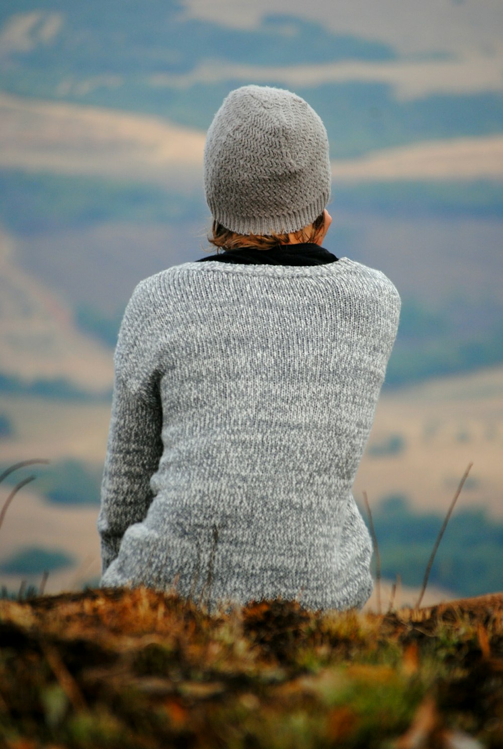 person in gray sweater sitting on green grass overlooking mountian