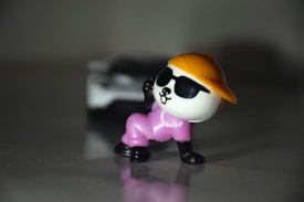 A small toy figure of a panda wearing a yellow cap, black sunglasses, and a pink outfit is posed in a playful stance. The figure is placed on a reflective surface with a blurred background.
