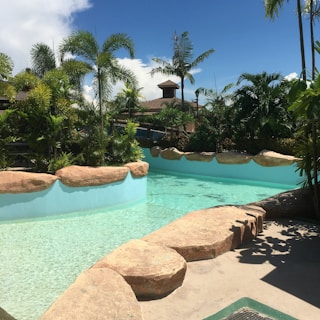A tropical swimming pool is surrounded by lush green palm trees and vegetation under a clear blue sky. Large rocks border the pool, adding a natural look to the setting. A small building with a wooden roof is visible in the background among the greenery.