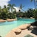 A tropical swimming pool is surrounded by lush green palm trees and vegetation under a clear blue sky. Large rocks border the pool, adding a natural look to the setting. A small building with a wooden roof is visible in the background among the greenery.