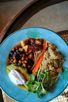 A colorful dish arranged in a blue bowl containing a variety of healthy ingredients. The dish includes quinoa, chickpeas, sliced carrots, sun-dried tomatoes, avocado, a dollop of white sauce, and fresh greens. The presentation is vibrant and balanced.