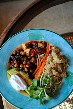A colorful dish arranged in a blue bowl containing a variety of healthy ingredients. The dish includes quinoa, chickpeas, sliced carrots, sun-dried tomatoes, avocado, a dollop of white sauce, and fresh greens. The presentation is vibrant and balanced.