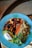 A colorful dish arranged in a blue bowl containing a variety of healthy ingredients. The dish includes quinoa, chickpeas, sliced carrots, sun-dried tomatoes, avocado, a dollop of white sauce, and fresh greens. The presentation is vibrant and balanced.