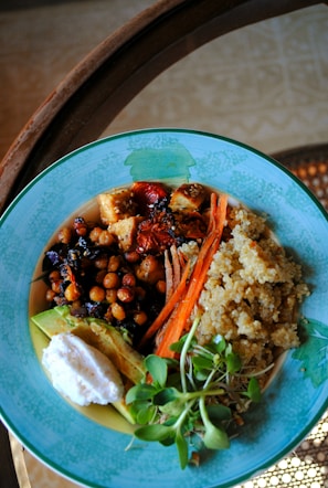 A colorful dish arranged in a blue bowl containing a variety of healthy ingredients. The dish includes quinoa, chickpeas, sliced carrots, sun-dried tomatoes, avocado, a dollop of white sauce, and fresh greens. The presentation is vibrant and balanced.