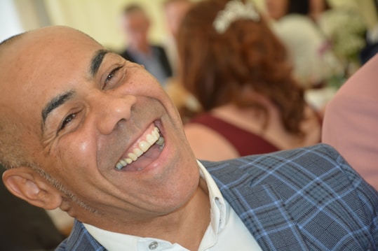 A person is laughing joyously, wearing a blue plaid jacket and a white shirt. The background is slightly blurred, featuring other people and soft lighting, suggesting a social gathering.