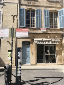 A historic building with pale stone walls and large blue shuttered windows. A variety of signs with directions and tourist information are visible at the base of the structure. The entrance includes a faded door and a window displaying signage, suggesting it might have been a bookstore or library.