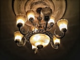 An ornate chandelier hangs from an intricately decorated ceiling, featuring multiple illuminated bulbs with an antique design. The warm glow of the bulbs contrasts with the darkened background, highlighting the fine details of the chandelier's metalwork and glass shades.