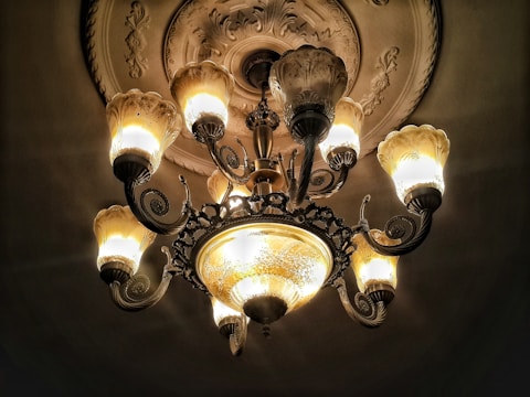 An ornate chandelier hangs from an intricately decorated ceiling, featuring multiple illuminated bulbs with an antique design. The warm glow of the bulbs contrasts with the darkened background, highlighting the fine details of the chandelier's metalwork and glass shades.