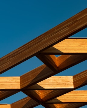 A close-up of rich wooden beams interlocking in a handcrafted timber frame structure against a clear blue sky.