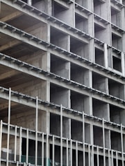 A building under construction with a visible concrete frame and multiple rectangular openings arranged in a grid pattern. The structure appears unfinished, showing bare concrete surfaces and metal rods.