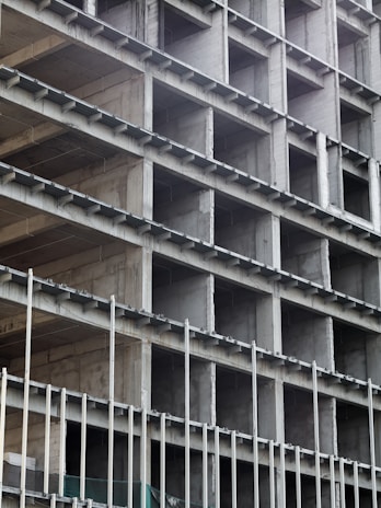A building under construction with a visible concrete frame and multiple rectangular openings arranged in a grid pattern. The structure appears unfinished, showing bare concrete surfaces and metal rods.
