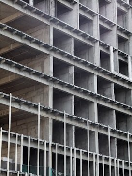 A building under construction with a visible concrete frame and multiple rectangular openings arranged in a grid pattern. The structure appears unfinished, showing bare concrete surfaces and metal rods.