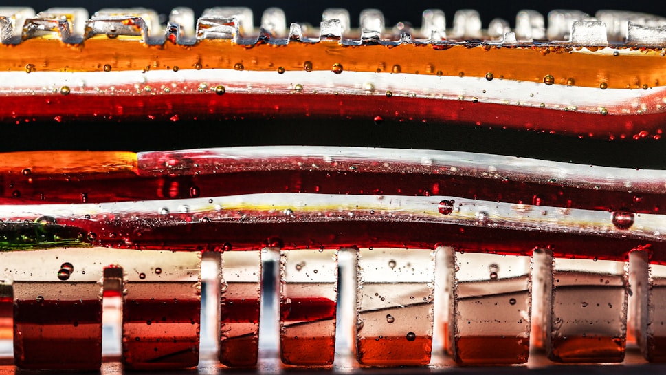 A close-up view of layered translucent materials with vivid colors including red, orange, and white. The layers are interspersed with small bubbles and are stacked in an abstract, artistic manner. The surface reflects light, creating a vibrant and visually dynamic image.