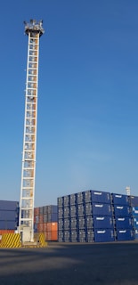 A tall metal tower with multiple lights stands near a large stack of shipping containers, which are primarily blue with some orange and black ones mixed in. The ground is paved, and the sky above is clear with a bright blue hue.