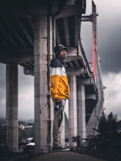 A person wearing a yellow and black jacket stands confidently under a large bridge, holding a camera. The atmosphere is moody with overcast skies and the backdrop features concrete structures and distant hills.