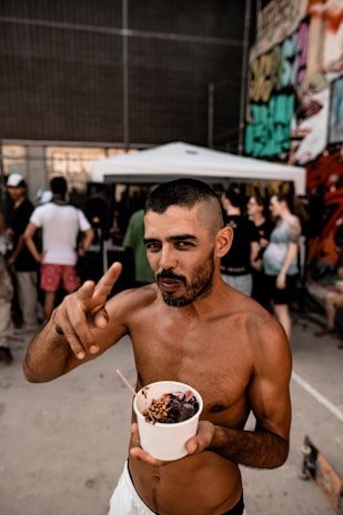 A shirtless man stands outdoors holding a cup filled with what appears to be acai topped with granola. He is making a gesture with his hand, and there are people and graffiti-covered walls in the background.