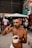 A shirtless man stands outdoors holding a cup filled with what appears to be acai topped with granola. He is making a gesture with his hand, and there are people and graffiti-covered walls in the background.