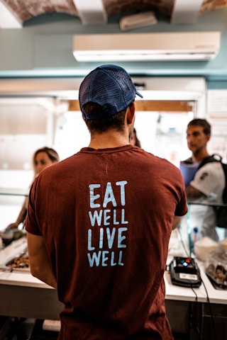 A person wearing a dark red T-shirt with the words 'EAT WELL LIVE WELL' printed in light blue stands at a counter. The person is facing away from the camera. The setting appears to be a casual indoor environment, possibly a food establishment, with two other people in conversation in the background. A point-of-sale terminal is visible on the counter.