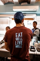 A person wearing a dark red T-shirt with the words 'EAT WELL LIVE WELL' printed in light blue stands at a counter. The person is facing away from the camera. The setting appears to be a casual indoor environment, possibly a food establishment, with two other people in conversation in the background. A point-of-sale terminal is visible on the counter.